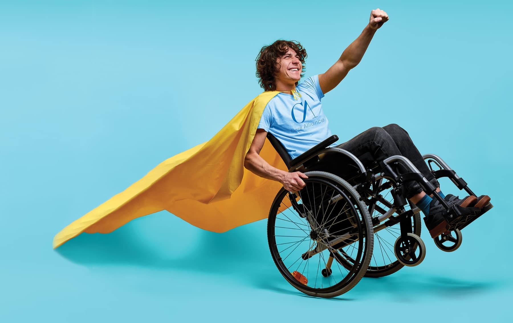 Smiling man in a wheelchair wearing a Care Alliance t-shirt and a yellow superhero cape, fist raised — the artwork on our wheelchair-accessible vans